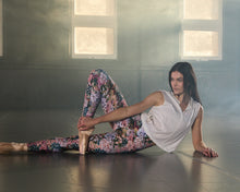 Load image into Gallery viewer, A woman in a white sleeveless top and liberty floral print leggings poses on the floor in a dimly lit studio with large windows.